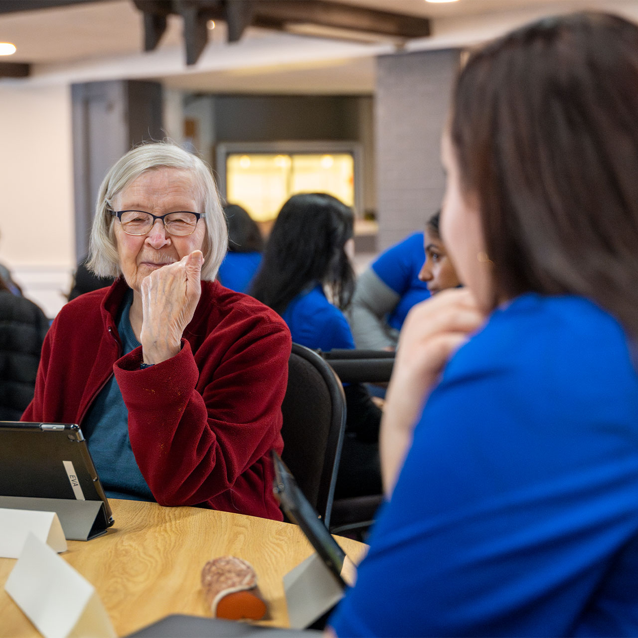 An elderly woman in a red jacket smiles while engaging with a young individual in a blue shirt