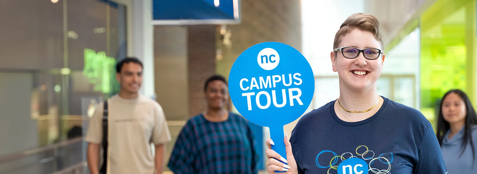 Women holding an NC Campus Tour sign while a tour group looks at her from behind.