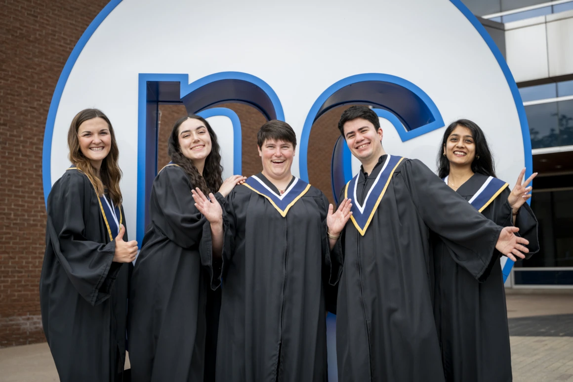 A group of graduates from NC’s Class of 2025 stand outside the Welland Campus in front of the NC button wearing graduation gowns.