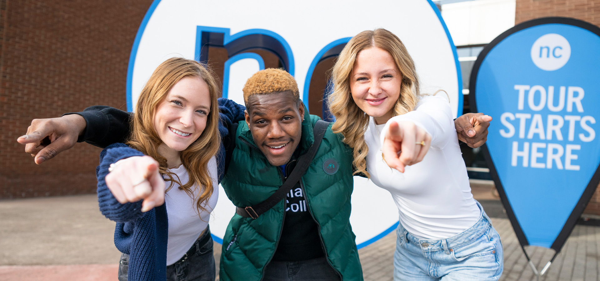 students standing outside smiling and pointing to the camera with a tour starts here sign behind them